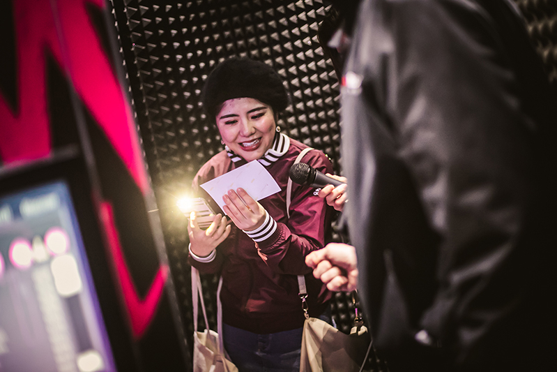 A woman holding a phone and a picture of herself inside the Pop-up Karaoke pod